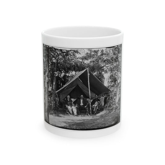 Black and white view of three seated men before a wall tent among tall trees, with laundry hung nearby.