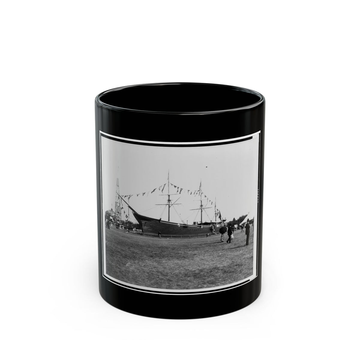 Black and white view of an old sailing ship on a grassy field, decorated with pennant flags and visitors nearby.