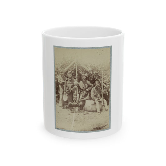 Sepia view of four soldiers in kepi caps resting under a leafy lean-to, one seated on a barrel with a rifle.