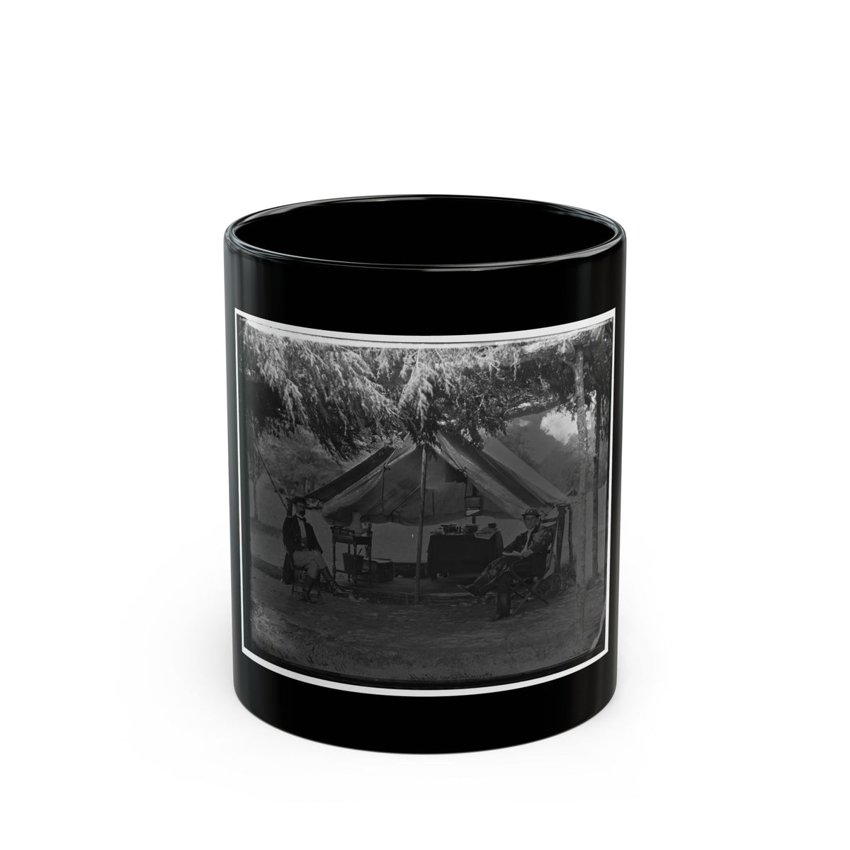 Black and white camp scene of two men seated outside a canvas tent under overhanging trees, with a camp table.