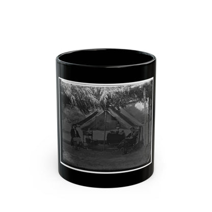 Black and white camp scene of two men seated outside a canvas tent under overhanging trees, with a camp table.