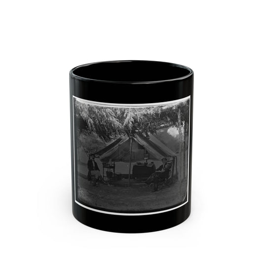 Black and white camp scene of two men seated outside a canvas tent under overhanging trees, with a camp table.