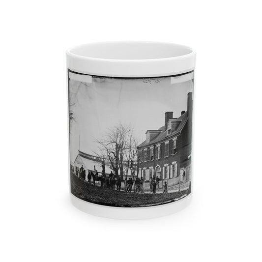 Black and white 19th-century view of uniformed soldiers and horses by a fence in front of a large brick house.