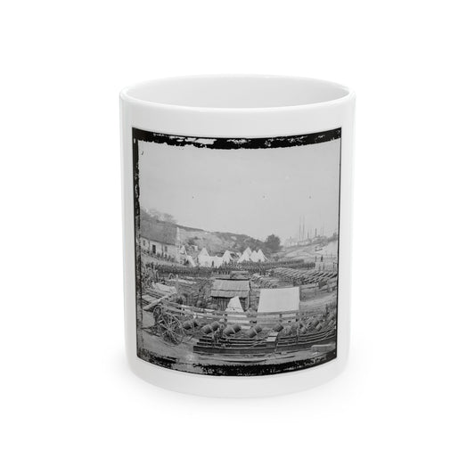 Black and white view of an army camp with rows of cannons, conical tents, and ships at the shore.