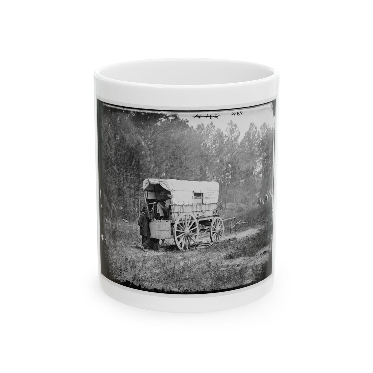 Black-and-white view of a canvas-covered supply wagon with spoked wheels on grass, a man in a long coat beside it.