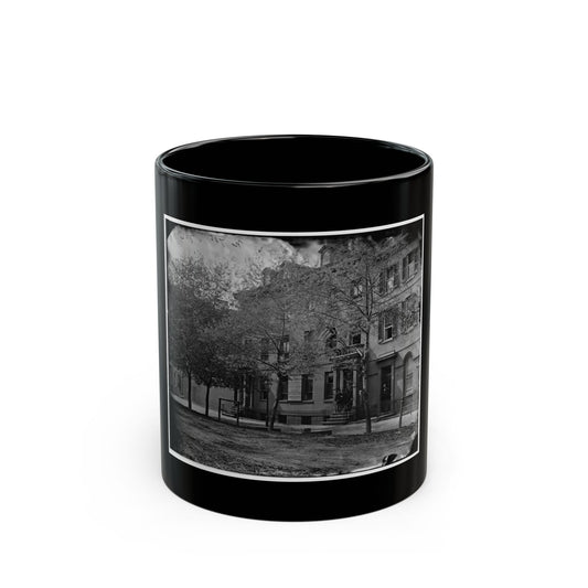 Black and white 1860s view of brick townhouses behind iron railings and bare street trees on a city street.