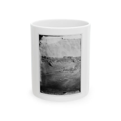 Black and white view of a ruined masonry fort with earthen ramparts, rubble, stacked barrels and a wooden pole.