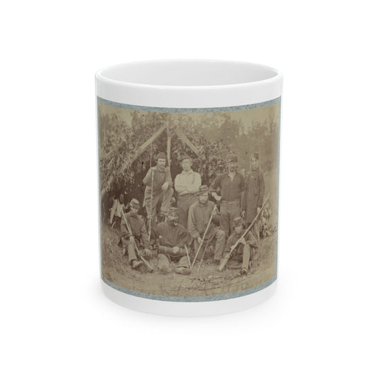 Sepia-toned print of seven uniformed soldiers with rifles in front of a leafy lean-to shelter, one seated with a pipe.