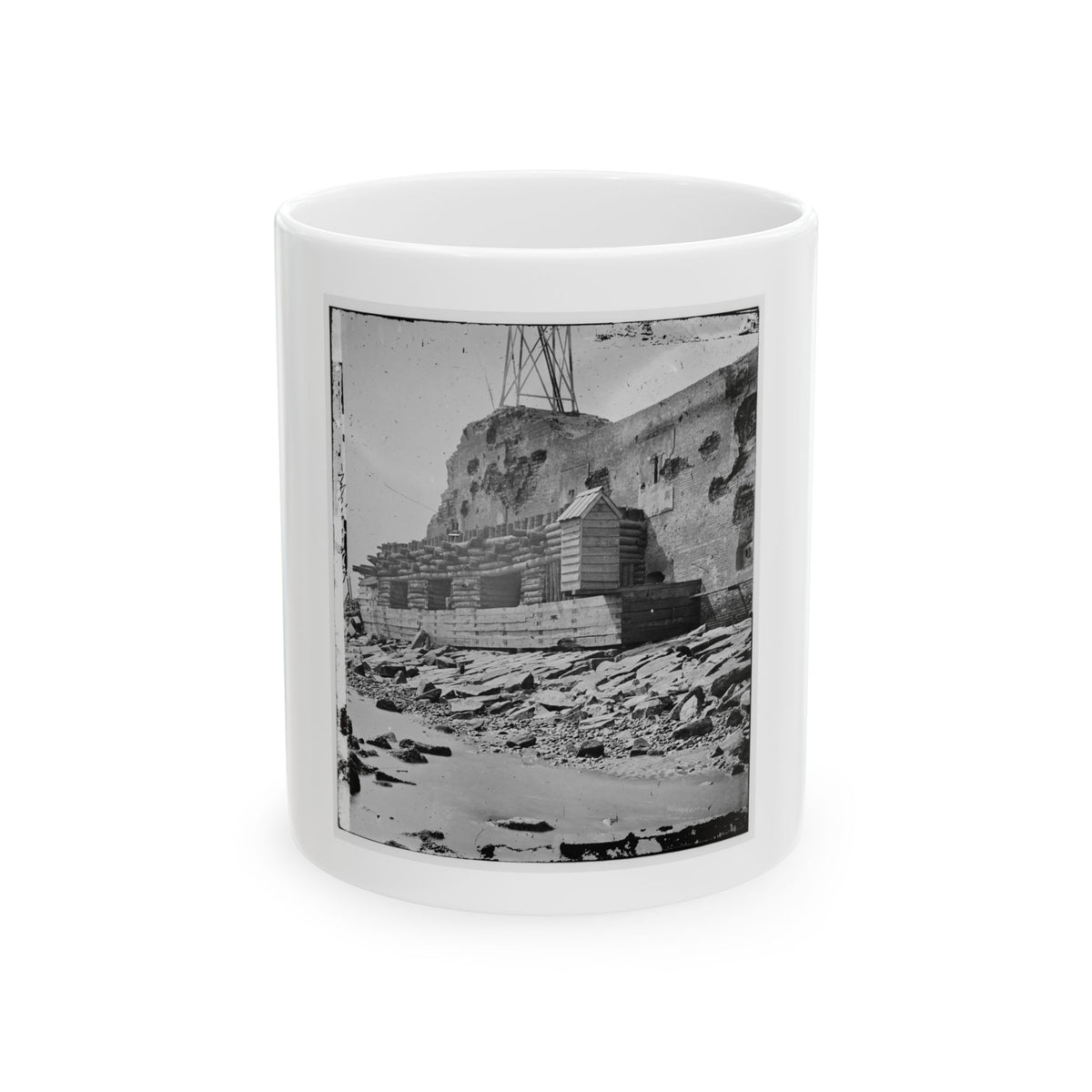 Black-and-white view of a damaged brick fort wall with timber revetment, a sentry box and rocks by the water.
