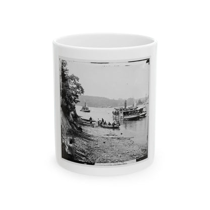 Black and white view of steamboats and a rowboat of seated men on a river shore beside a wooded bank.