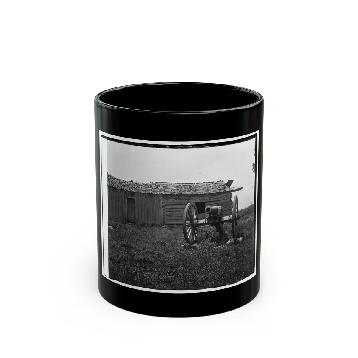 Black-and-white view of a field cannon on a wooden carriage beside a weathered log cabin in tall grass.
