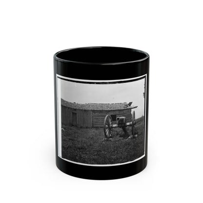 Black-and-white view of a field cannon on a wooden carriage beside a weathered log cabin in tall grass.