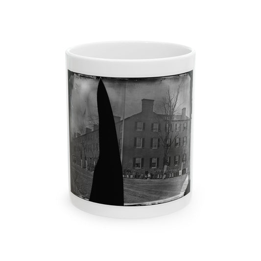 Black and white view of a long brick barracks with many windows, soldiers standing in line before it under bare trees.