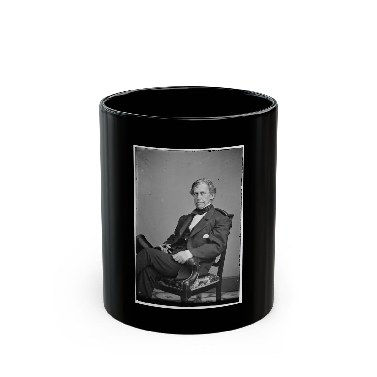 Black and white studio portrait of a seated man in a dark suit and cravat, legs crossed in a carved armchair.