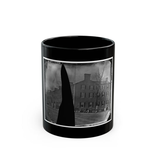 Black and white view of a long brick barracks with many windows, soldiers standing in line before it under bare trees.