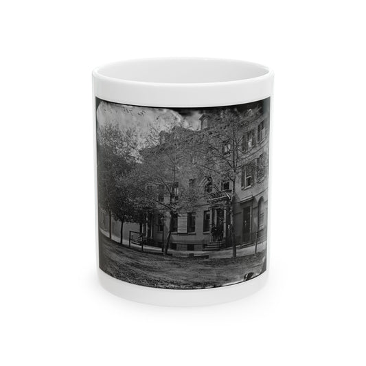 Black and white 1860s view of brick townhouses behind iron railings and bare street trees on a city street.