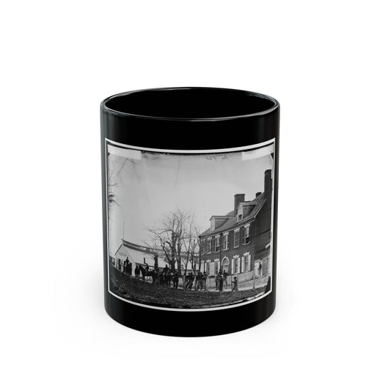 Black and white 19th-century view of uniformed soldiers and horses by a fence in front of a large brick house.
