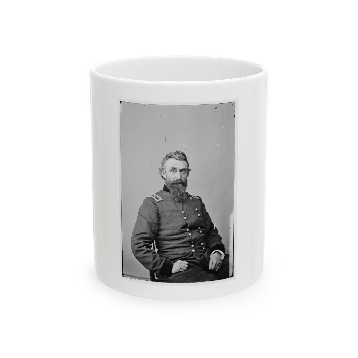 Black and white studio portrait of a bearded man in a double-breasted military coat, seated against a plain grey backdrop.