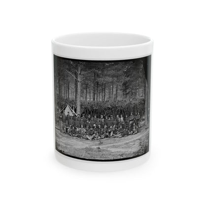 Black and white view of a large group of uniformed soldiers posed outdoors among pine trees beside a tent.
