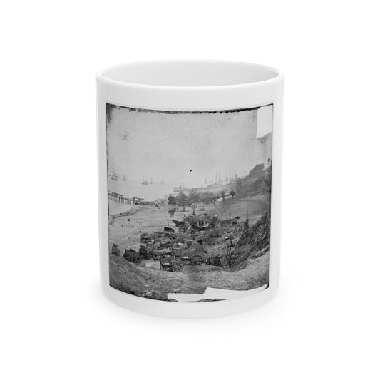 Black and white view of many supply wagons parked on a sandy shore with sailing ships and a pier in the harbor.