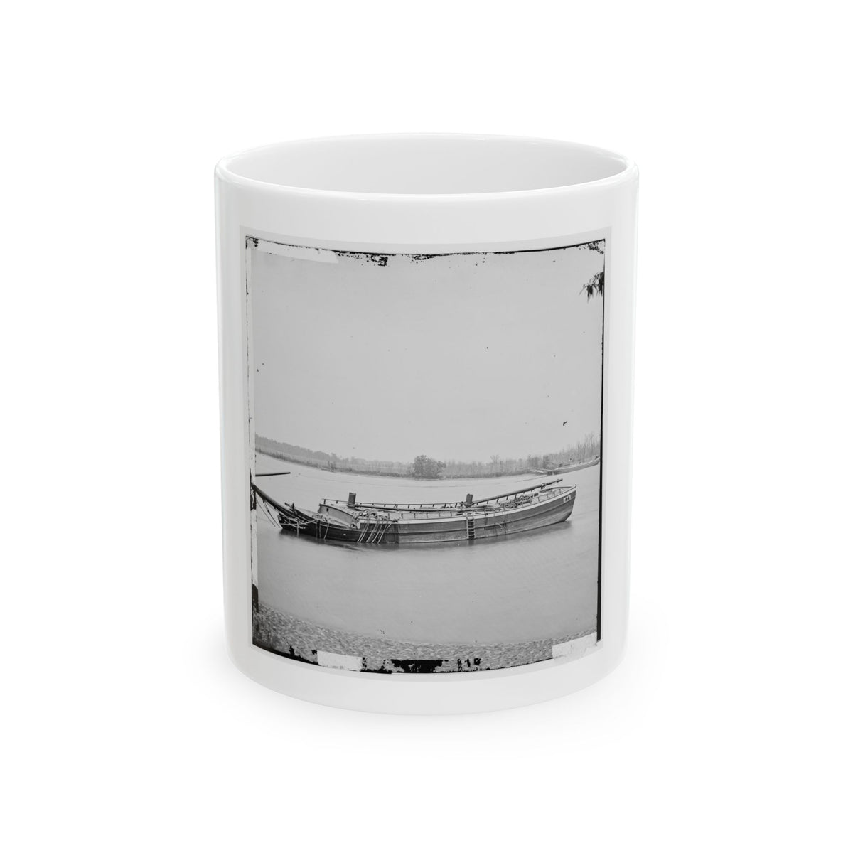 Black and white view of a wooden riverboat with long spars moored on a calm river, with trees on the far shore.